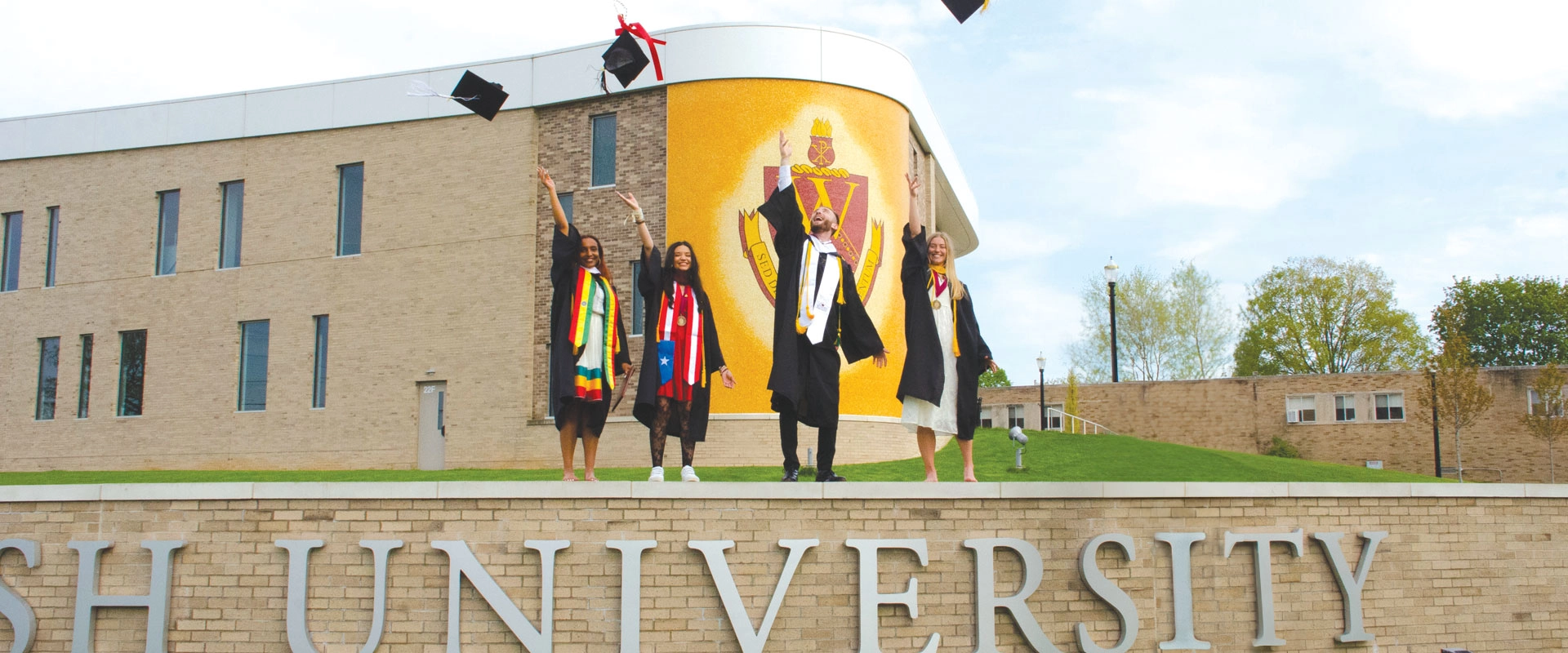 photo of four students standing in front of the Marlene and Joe Toot Global Learning Center Walsh crest mosaic tossing graduation caps in the air
