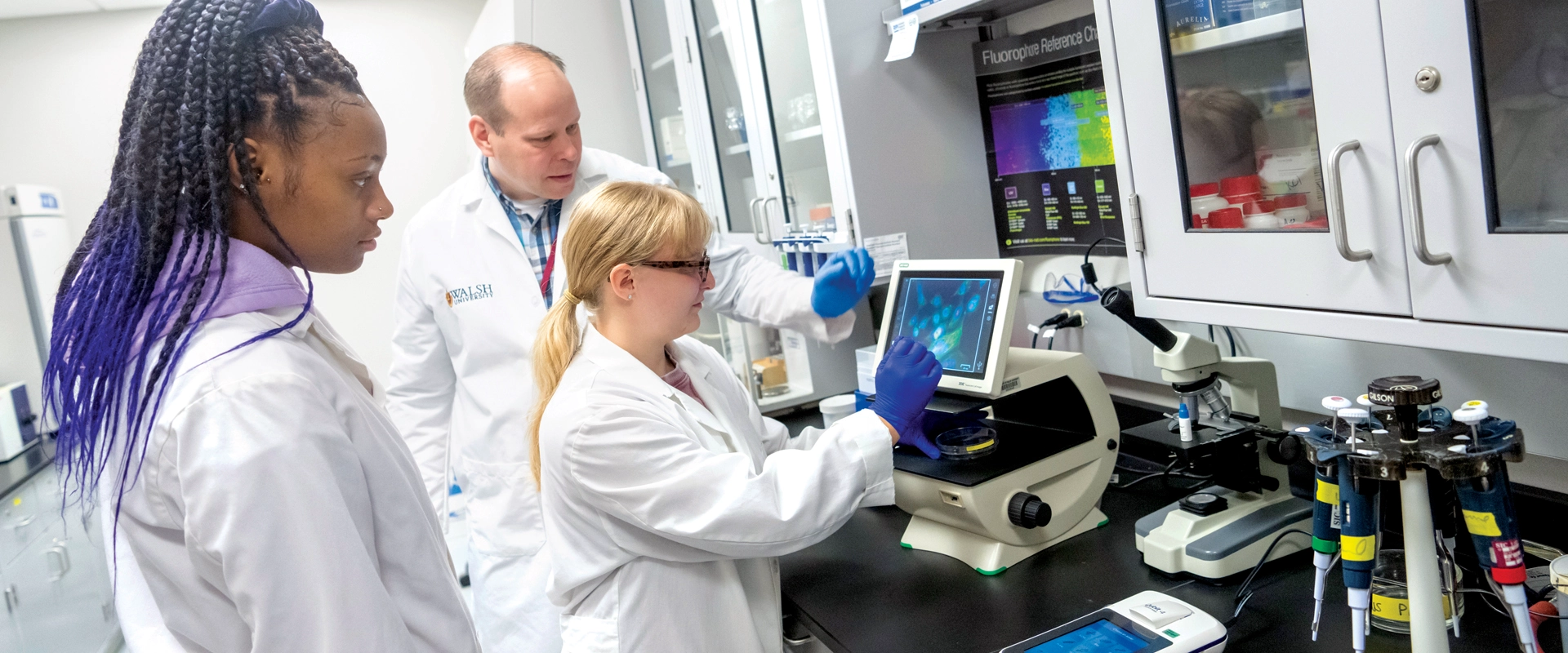 photo of two students in the biology lab looking at an enlarged image from a microscope while the instructor looks on