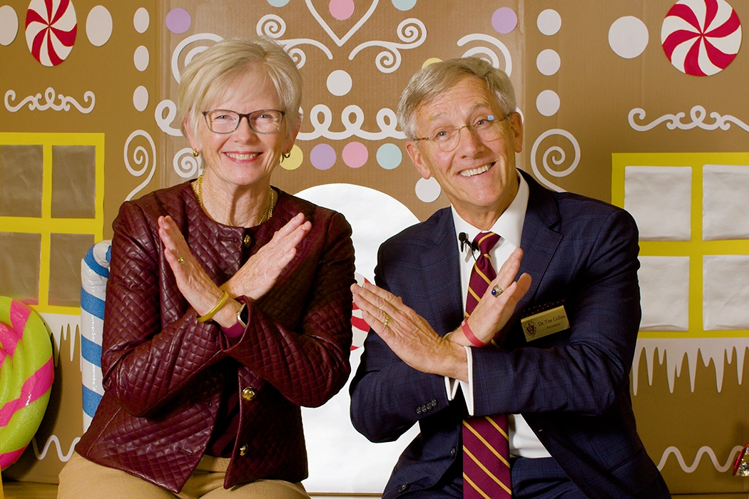 Photo of Walsh University President Dr. Tim and Drenda Collins seated in front of a gingerbread house scene smiling and presenting a Swords Up gesture (arms crossed in front of body)